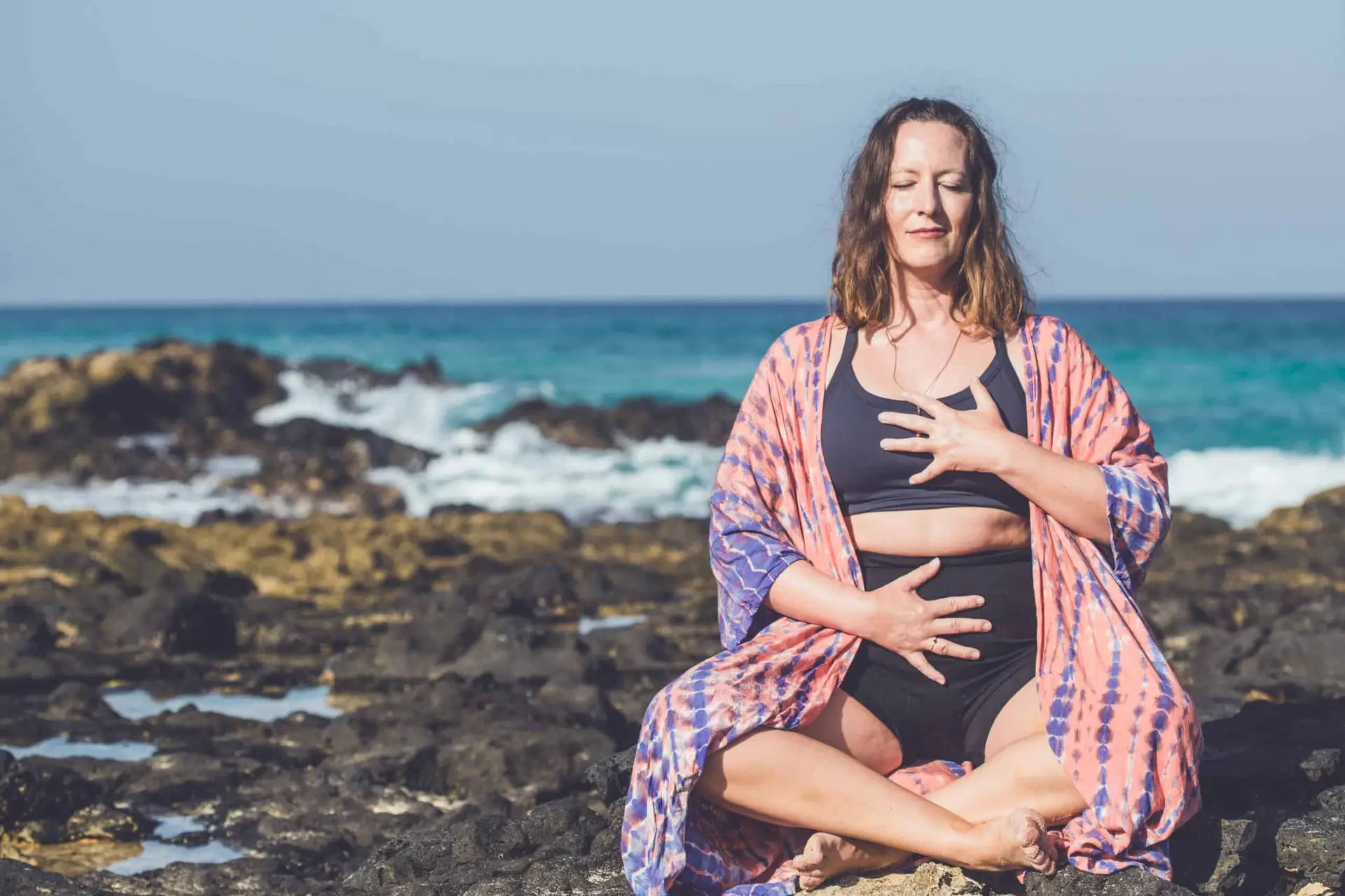 Sandra sitzt am Meer mit einer Hand auf der Brust und einer Hand auf dem Bauch
